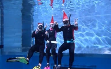 Three freedivers underwater in wetsuits and Santa hats, each giving a peace sign.